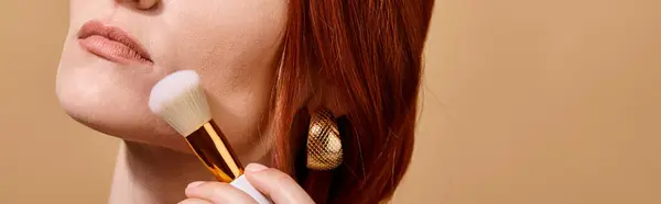 Redhead woman in gold earring applying face foundation with makeup brush on beige backdrop, banner — Stock Photo