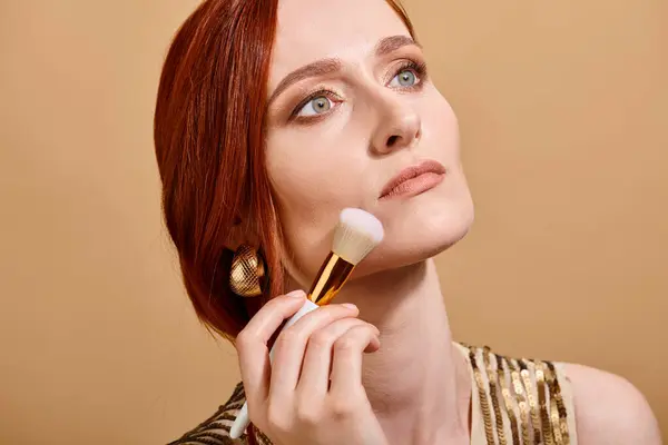 Rossa donna in oro orecchino applicando fondotinta viso con pennello trucco su sfondo beige — Foto stock
