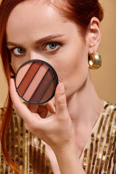 Redhead woman covering her face with eye shadow palette on beige background, makeup concept — Stock Photo