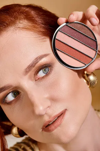 Close up of redhead woman holding eye shadow palette near face on beige background, makeup concept — Stock Photo
