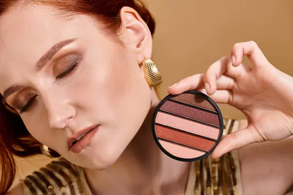 Nahaufnahme einer rothaarigen Frau mit Lidschattenpalette in Gesichtsnähe auf beigem Hintergrund, Make-up-Konzept — Stockfoto