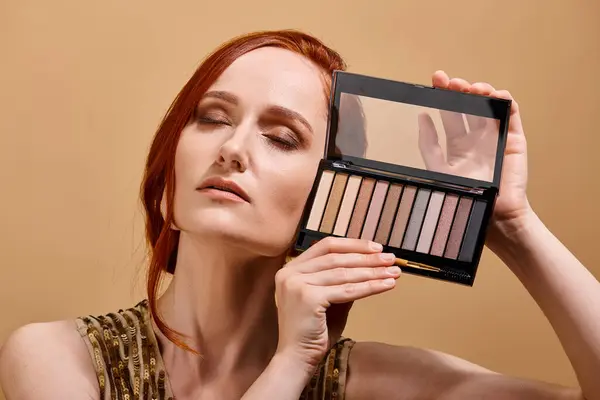 Redhead woman holding eye shadow palette near face on beige background, makeup advertisement — Stock Photo