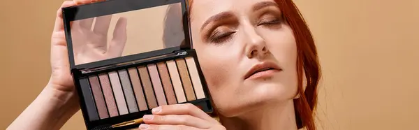 Rousse femme tenant ombre à paupières palette près du visage sur fond beige, bannière publicitaire maquillage — Photo de stock