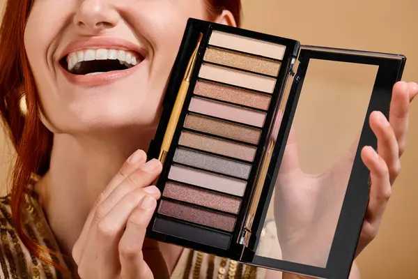 Cropped view of happy woman holding eye shadow palette on beige background, makeup advertisement — Stock Photo