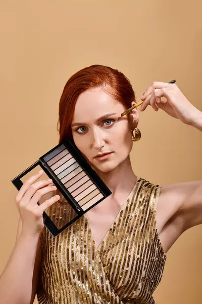 Redhead woman applying eye shadow and holding palette on beige background, pastel colors — Stock Photo