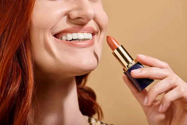 Cropped view of redhead woman smiling and applying nude lipstick on beige background, makeup product — Stock Photo
