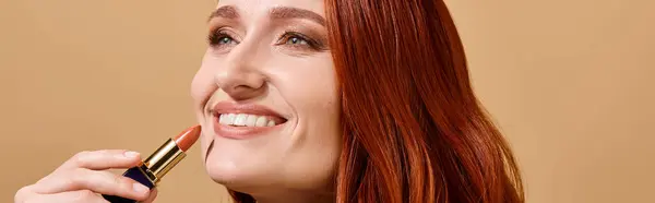 Fröhliche und rothaarige Frau lächelt und trägt Nude-Lippenstift auf beigem Hintergrund auf, Make-up-Banner — Stockfoto
