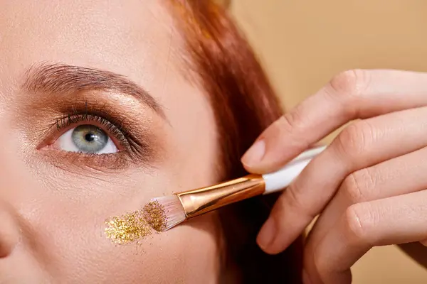 Close up of redhead woman applying sparkling glitter on cheek with makeup brush on beige backdrop — Stock Photo