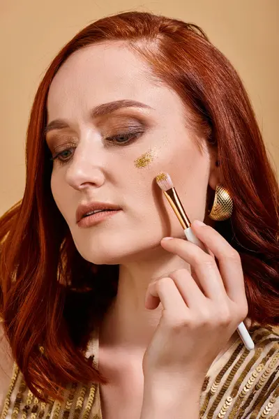 Radiant and redhead woman applying sparkling glitter on cheek with makeup brush on beige background — Stock Photo