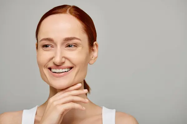 Cheerful and redhead woman in her 30s posing in white tank top on grey background, genuine smile — Stock Photo