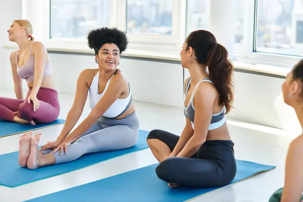 Felices amigas desnudas y multiculturales charlando mientras están sentadas en colchonetas de yoga antes de clase - foto de stock