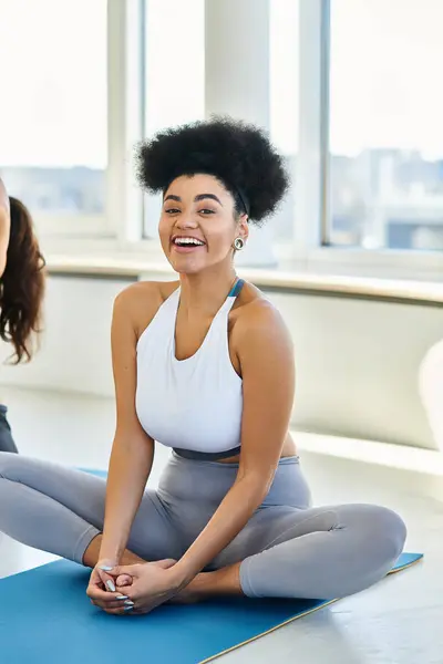 Fröhliche Afroamerikanerin in Sportbekleidung sitzt auf Matte und genießt Yoga-Kurs drinnen — Stockfoto