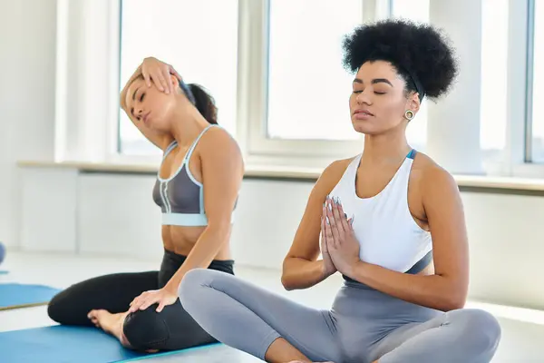 Enfoque en la mujer afroamericana en ropa deportiva meditando en postura de yoga en la estera cerca de amigo borrosa - foto de stock