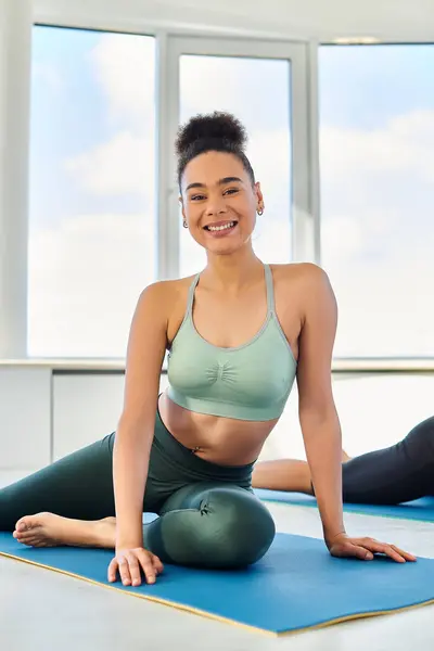 Feliz africana americana mujer en sportswear estiramiento en yoga mat cerca amigo en borrosa fondo - foto de stock