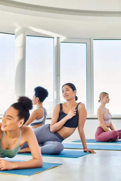 Se centran en sonreír asiático en ropa deportiva sentado en la alfombra de fitness cerca de su interracial amigas - foto de stock