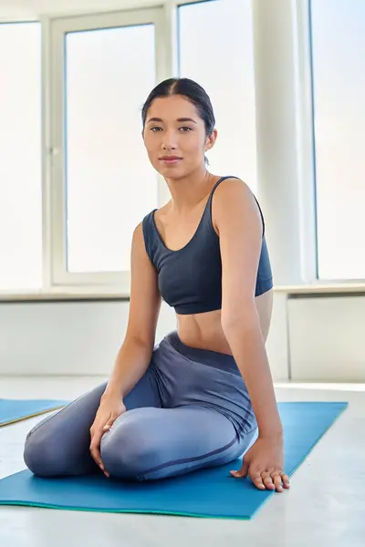 Joven y morena asiática mujer en su 20s sentado en yoga mat y mirando a cámara, deportivo - foto de stock