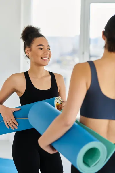 Mulheres felizes multiétnicas e jovens na casa dos 20 anos de pé com tapetes de ioga e bate-papo, pós-treino — Fotografia de Stock