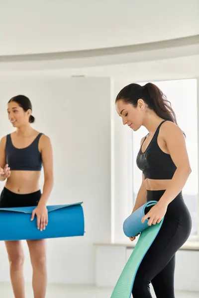 Brunette jeune femme en vêtements de sport debout avec tapis de yoga à côté de flou asiatique ami en studio — Photo de stock