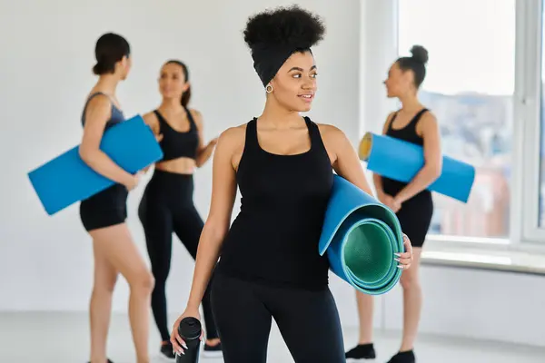 Enfoque en la mujer afroamericana feliz de pie con tapete de fitness y botella de deportes en el estudio de yoga - foto de stock