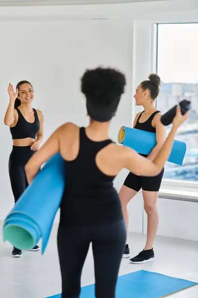 Mujer morena feliz agitando las manos a su amigo afroamericano con colchoneta de fitness en el estudio de yoga - foto de stock