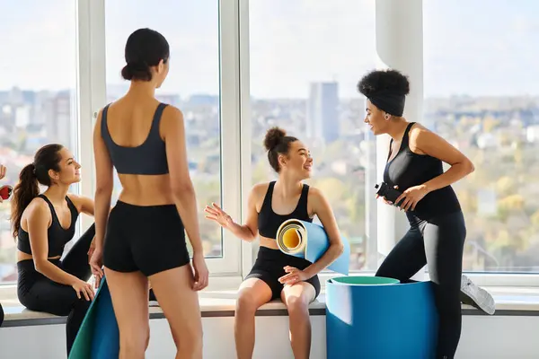 Feliz multicultural de las mujeres jóvenes en los años 20 charlando mientras se sienta al lado de la ventana en el estudio de pilates - foto de stock