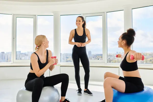 Happy multicultural friends exercising with dumbbells on fitness balls near female trainer — Stock Photo