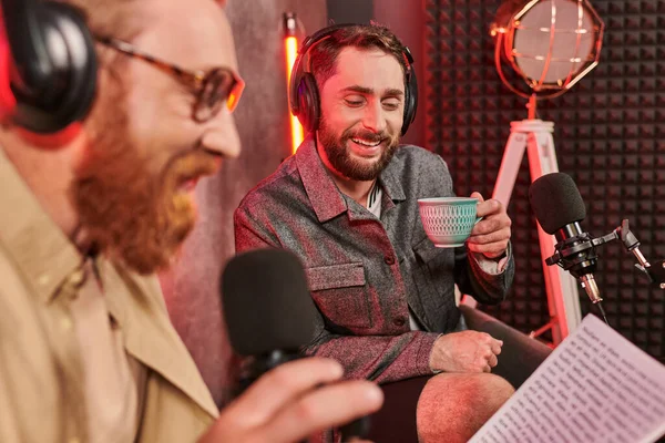 Dos hombres guapos barbudos en atuendos casuales con auriculares hablando durante el podcast, taza de café - foto de stock