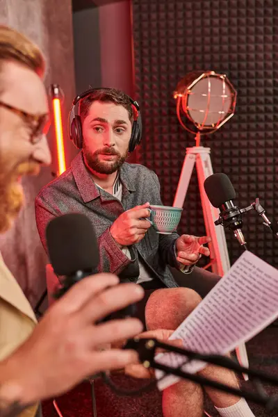 Two bearded handsome men in casual attires with headphones talking during podcast, coffee cup — Stock Photo