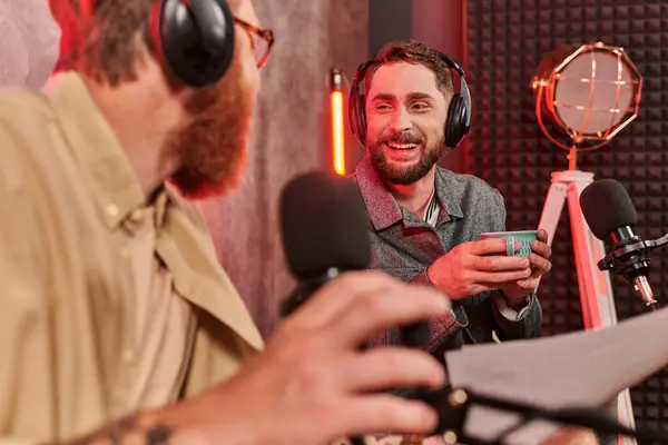 Two bearded handsome men in casual attires with headphones talking during podcast, coffee cup — Stock Photo