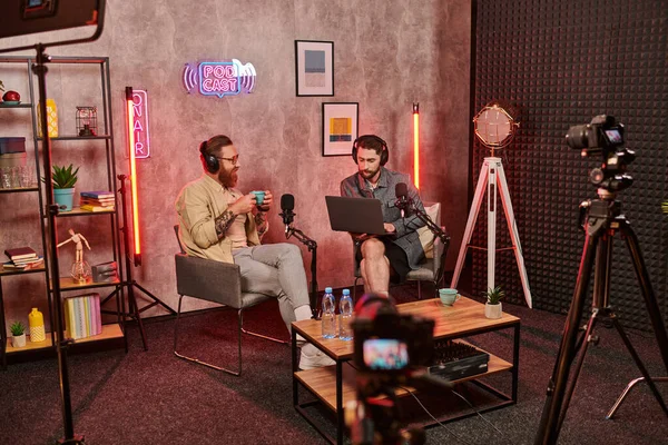 Hombres barbudos de buen aspecto con auriculares bebiendo café y hablando activamente durante el podcast - foto de stock