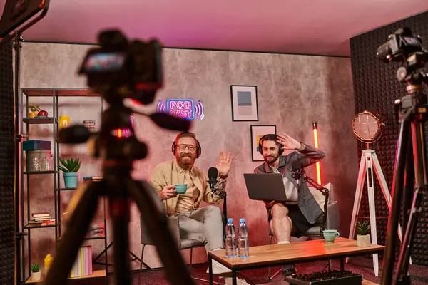 Beaux hommes barbus avec écouteurs buvant du café et parlant pendant le podcast, agitant les mains — Photo de stock