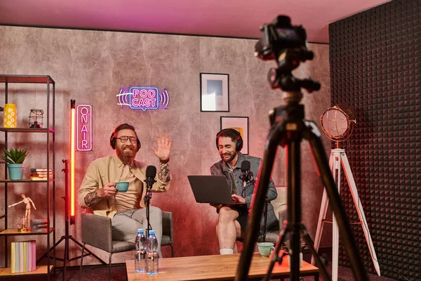 Hombres barbudos guapos con auriculares bebiendo café y hablando durante el podcast, agitando las manos - foto de stock