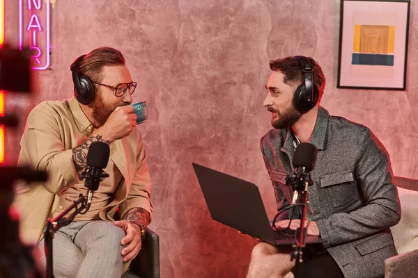 Gut aussehende bärtige Männer mit Kopfhörern, die Kaffee trinken und sich während des Podcasts aktiv unterhalten — Stockfoto