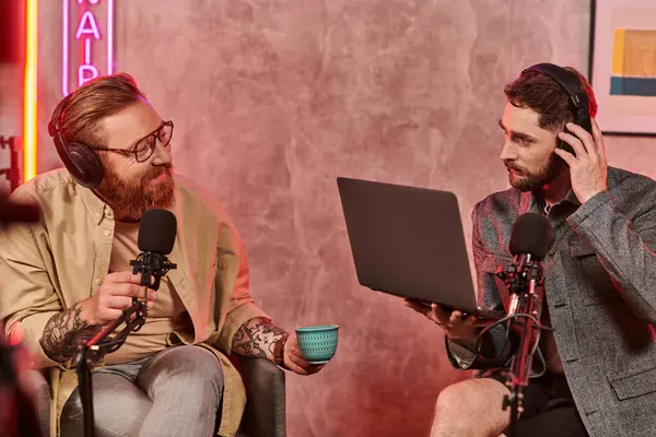 Hombres guapos en atuendos casuales posando con café y portátil durante su discusión en podcast - foto de stock
