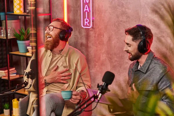 Hombres guapos en atuendos casuales posando con café y portátil durante su discusión en podcast - foto de stock