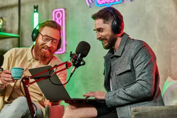 Hombres alegres atractivos en atuendos casuales con café y computadora portátil durante su discusión en podcast - foto de stock