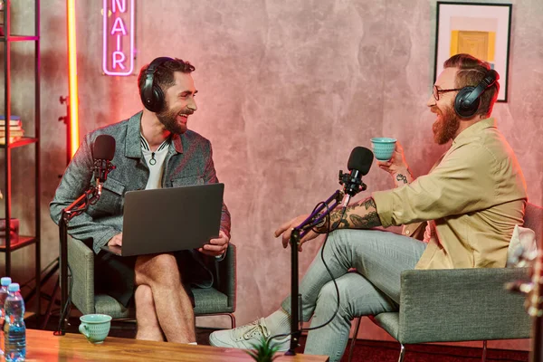 Fröhliche Männer in bequemen Anzügen mit Kaffee und Laptop während ihrer Podcast-Diskussion — Stockfoto
