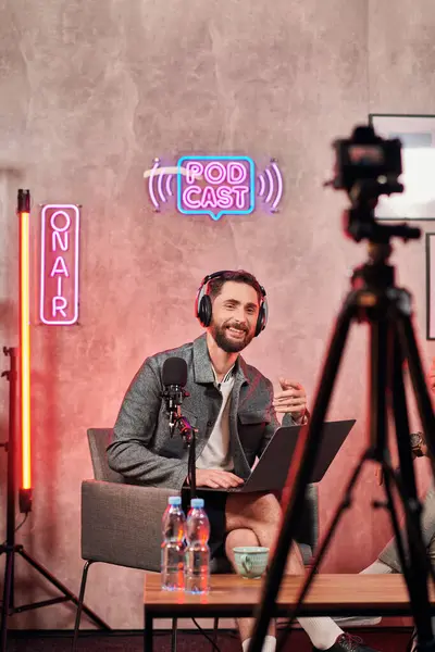 Hombre barbudo alegre en traje cómodo casual sentado con el ordenador portátil durante el podcast mirando a la cámara - foto de stock