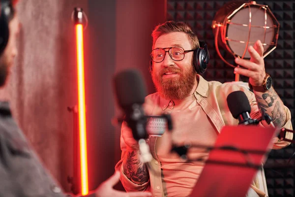 Cheerful handsome man with red beard with headphones in casual clothes talking during podcast — Stock Photo