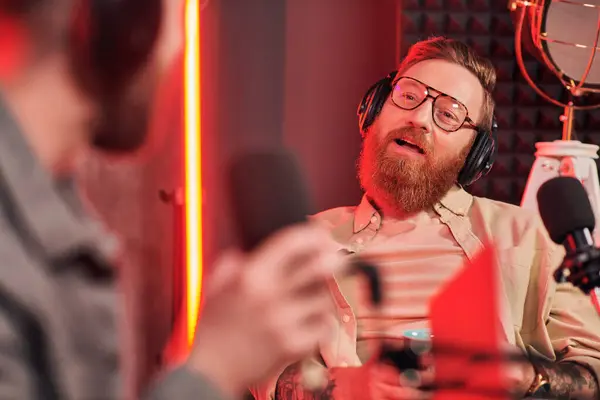 Entrevistador barbudo con gafas hablando con su invitado borroso durante su entrevista en podcast - foto de stock