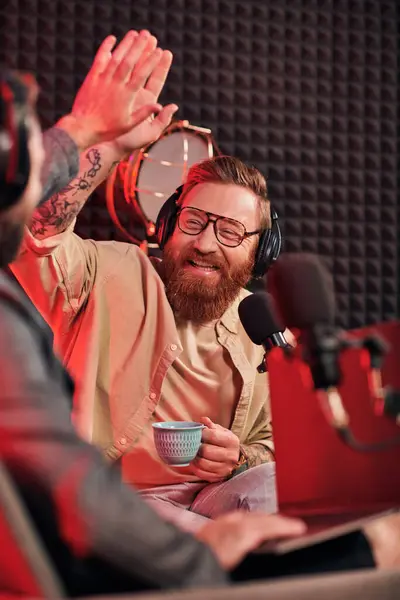 Entrevistador barbudo con gafas haciendo cinco altos con su invitado borroso durante su podcast - foto de stock