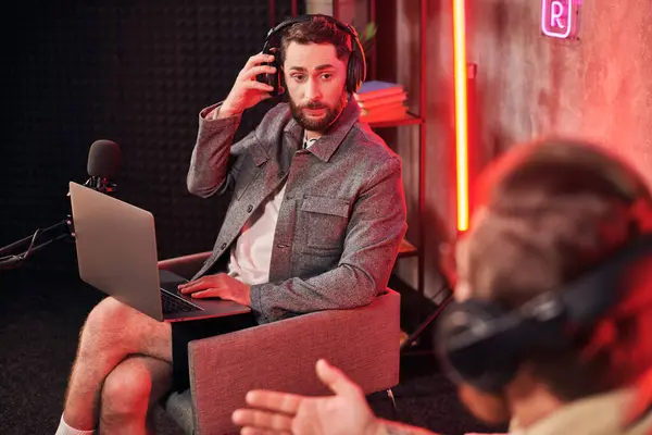 Bel homme barbu en tenue décontractée confortable assis avec ordinateur portable en studio à côté de son podcaster — Photo de stock