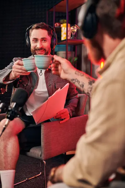 Весёлый бородатый мужчина с кофейной чашкой в руке смотрит на бумажную работу рядом со своим подкастером — стоковое фото
