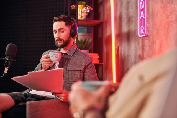 Hombre barbudo bien parecido con taza de café en la mano mirando el papeleo al lado de su podcaster - foto de stock
