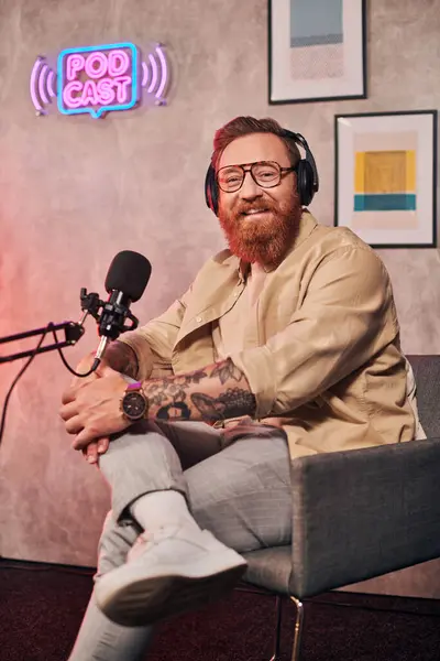 Joyeux bel homme aux cheveux roux et barbe avec écouteurs regardant la caméra pendant le podcast — Photo de stock