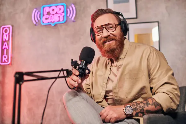 Allegro bell'uomo con i capelli rossi e la barba con le cuffie guardando la fotocamera durante il podcast — Foto stock