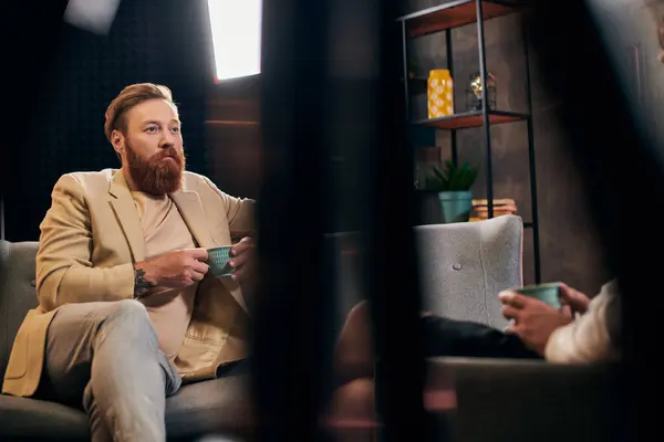 Bel homme barbu aux cheveux roux dans des vêtements élégants assis à côté de son intervieweur en studio — Photo de stock