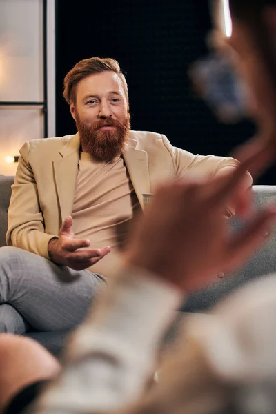 Hombre barbudo de buen aspecto con el pelo rojo en ropa elegante sentado al lado de su entrevistador en el estudio - foto de stock