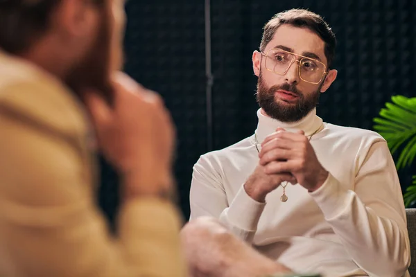 Bel homme barbu avec des lunettes dans une tenue élégante assis et regardant son intervieweur — Photo de stock