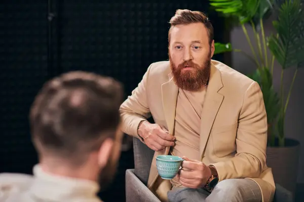 Hombre de pelo rojo bien parecido con taza de café sentado durante la discusión junto a su entrevistador - foto de stock